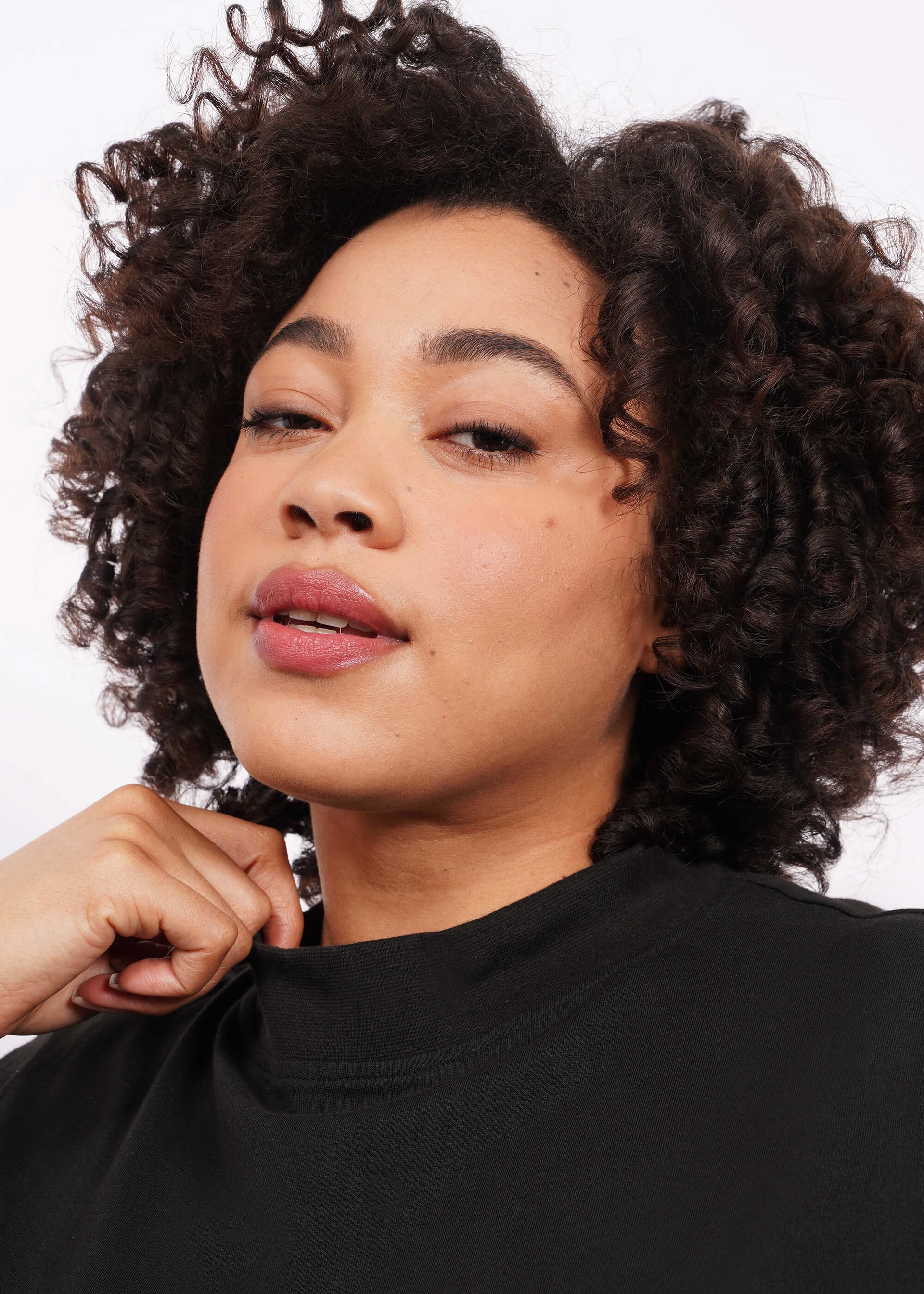 Close up view of a person with short curly dark hair against a white background, wearing The Essential Mock Neck Tank in black with one hand touching the shirt's collar and looking directly at the camera.
