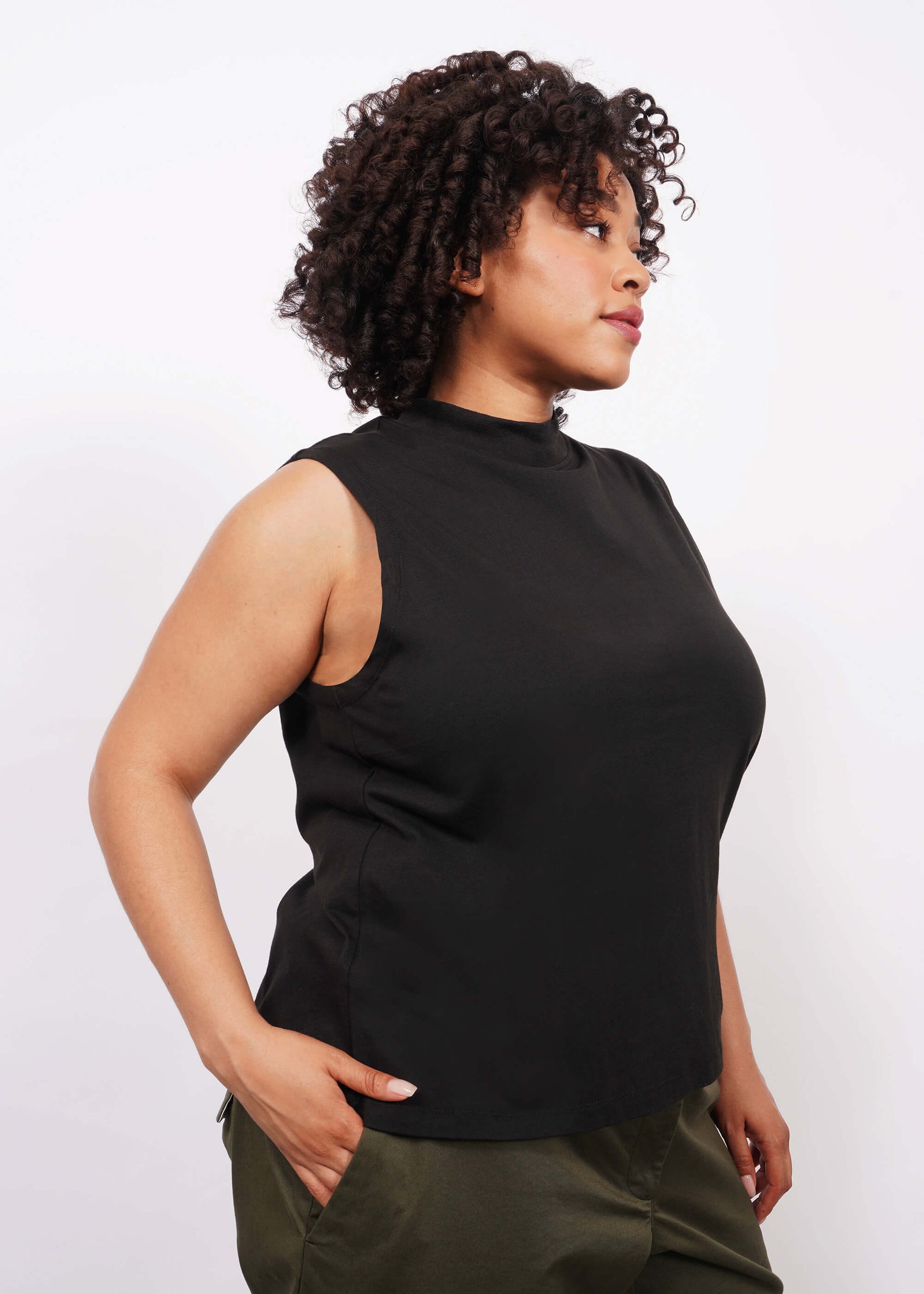 Side view of a person with short curly dark hair standing against a white background, wearing The Essential Mock Neck Tank and dark green pants, with one hand in the pant pocket, and looking to the side.