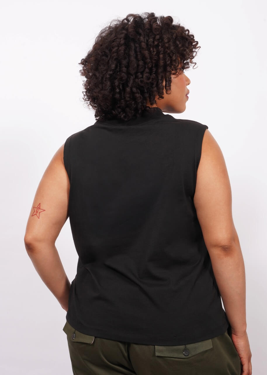 Back, waist up view of a person with short curly dark hair standing against a white background, wearing The Essential Mock Neck Tank and dark green pants, with one hand in the pant pocket.