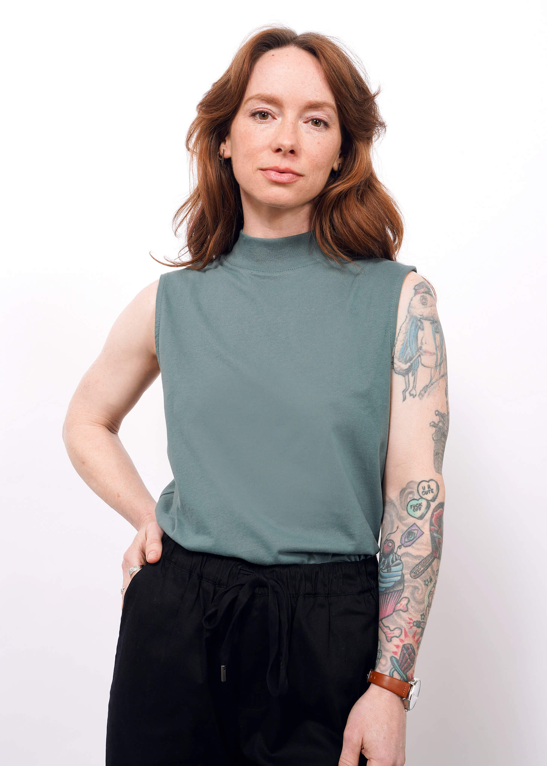 A woman with long brown hair and visible tattoos on her left arm stands confidently against a plain white background, wearing The Essential Mock Neck Tank in green paired with black pants.