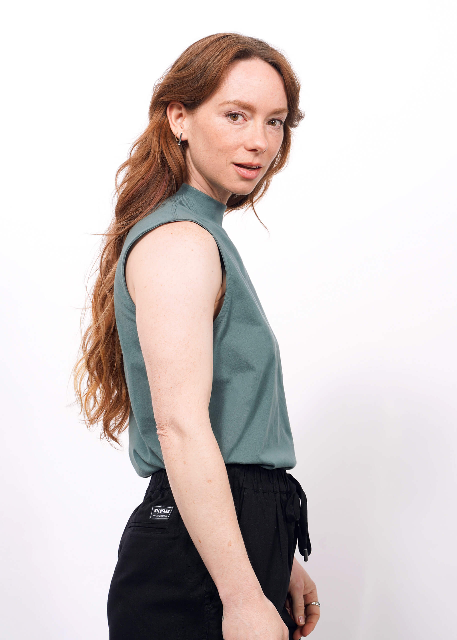 A woman with long, wavy reddish-brown hair stands sideways, facing the camera. She wears The Essential Mock Neck Tank in green and black drawstring pants against a plain white background.