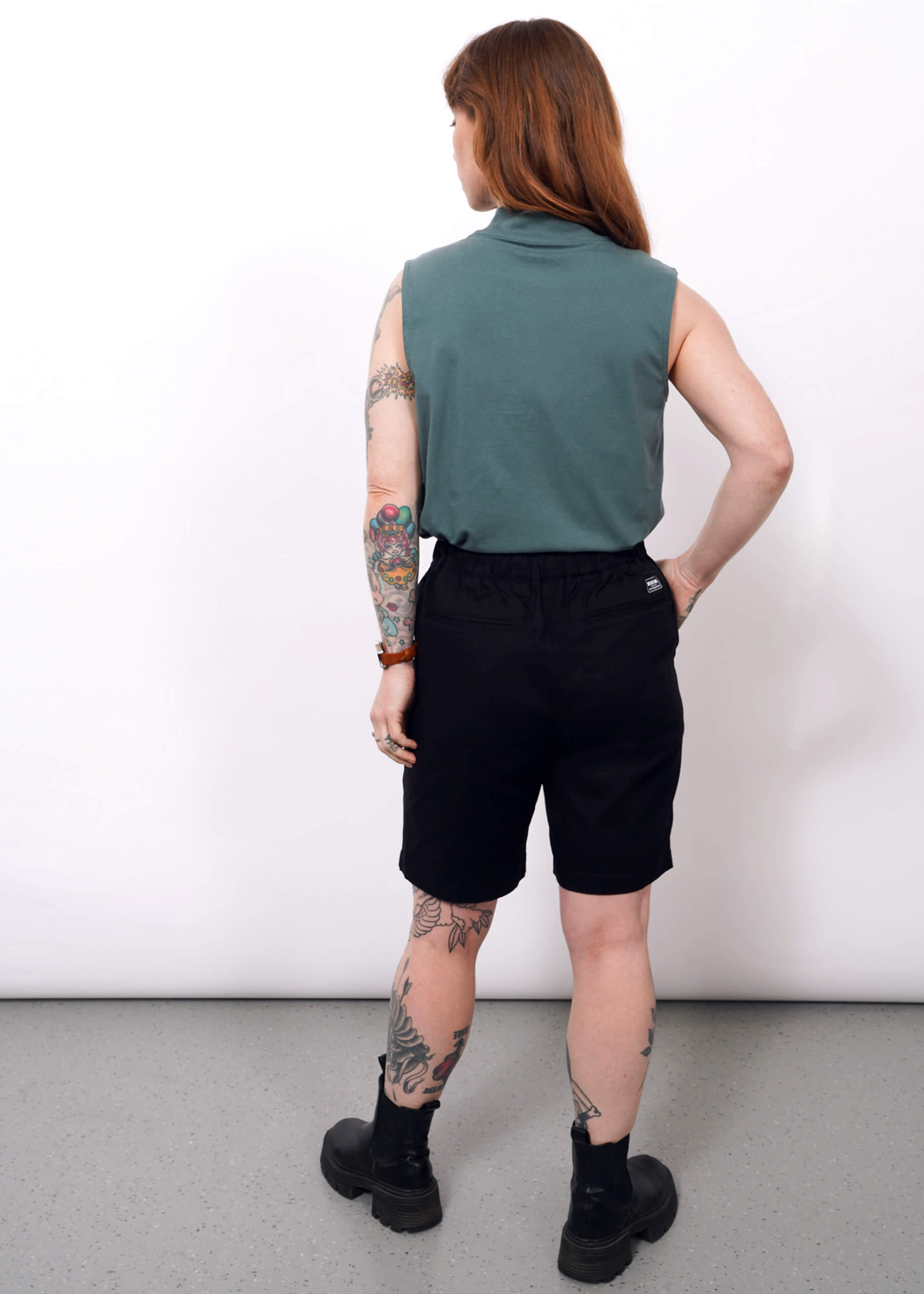 A person with long hair and visible tattoos wears The Essential Mock Neck Tank in green, paired with black shorts and black shoes, standing with their back to the camera against a plain white background.