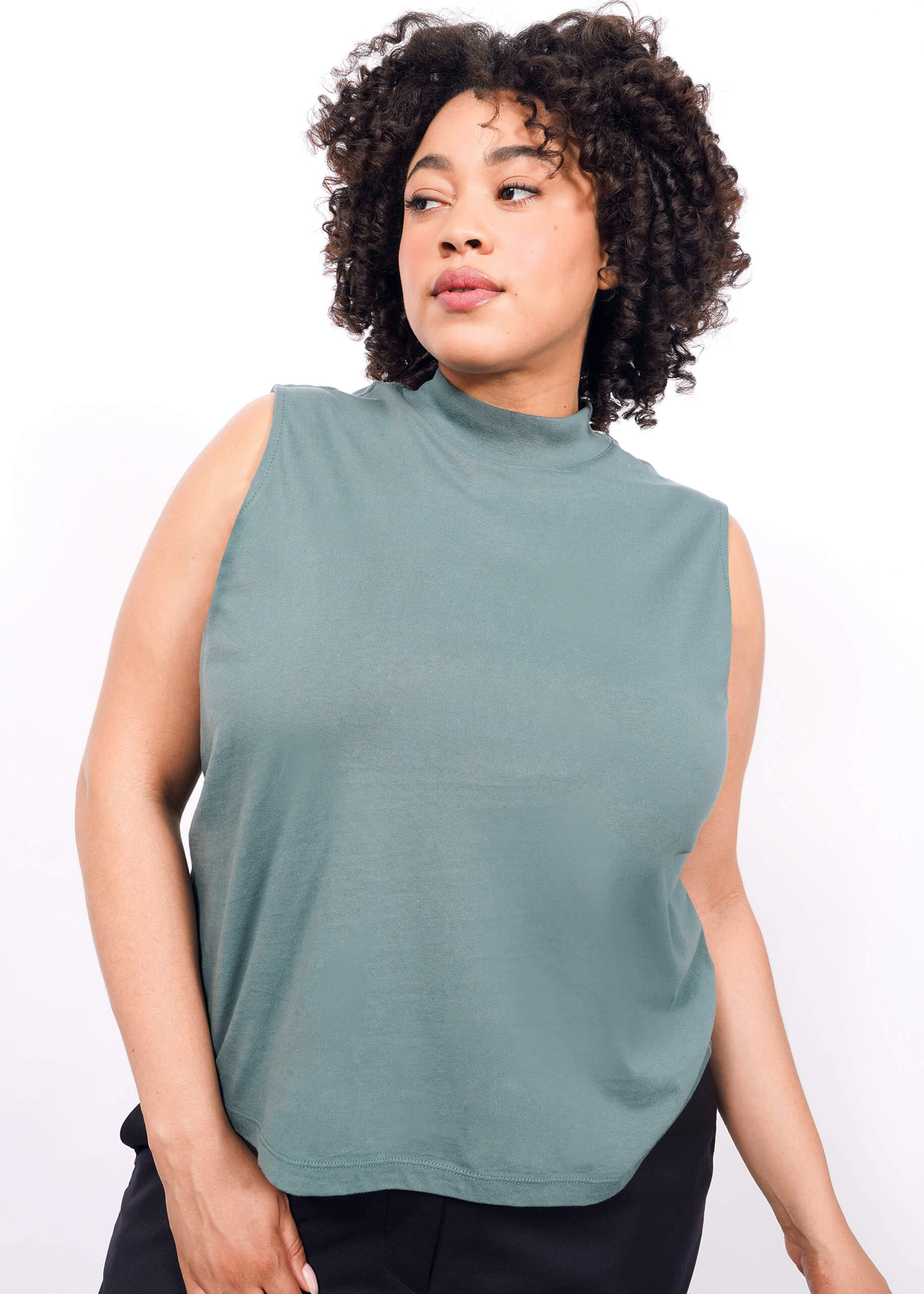 A woman with curly hair models The Essential Mock Neck Tank in sage green, paired with black pants. She stands against a white background, looking calm—this relaxed-fit tank is perfect for layering with any outfit.
