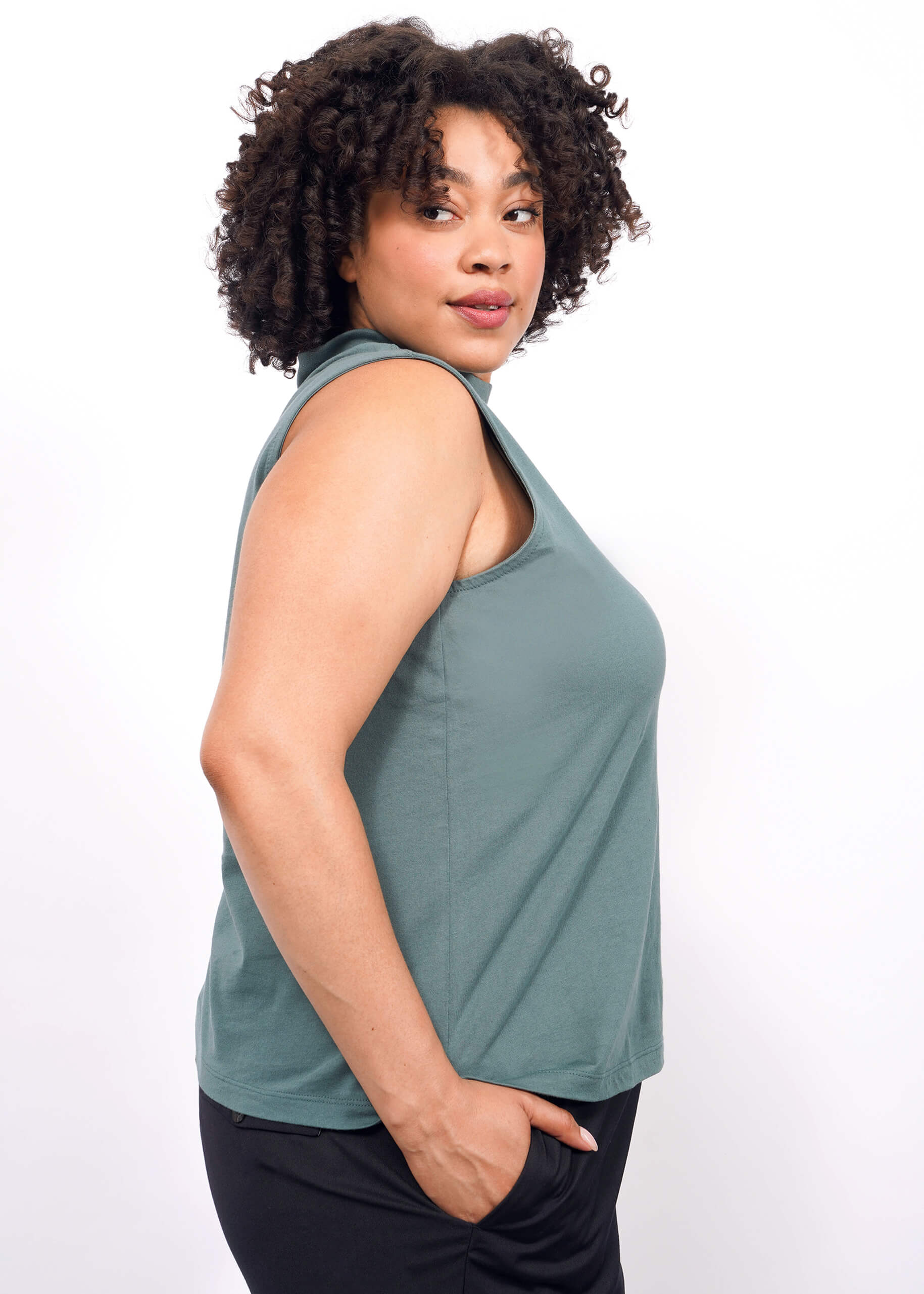 A woman with curly hair wears The Essential Mock Neck Tank in green and black pants, standing in profile against a white background and looking over her shoulder with a slight smile.