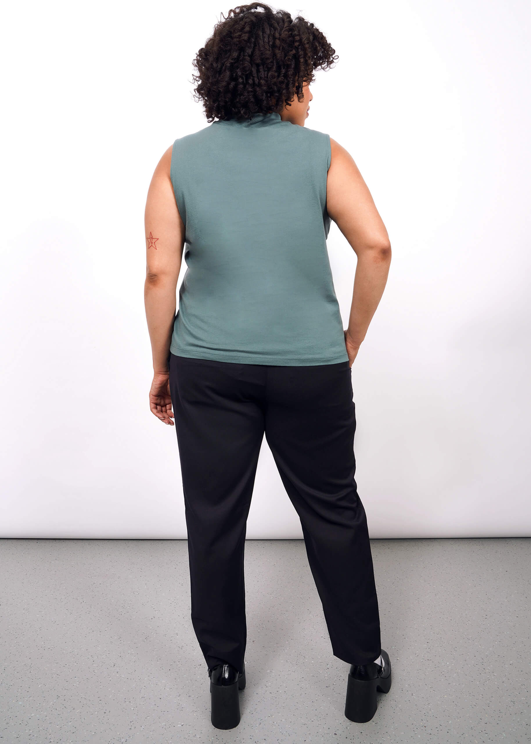 A person with curly hair, wearing The Essential Mock Neck Tank in green, black pants, and black shoes, stands facing away from the camera against a plain white background.