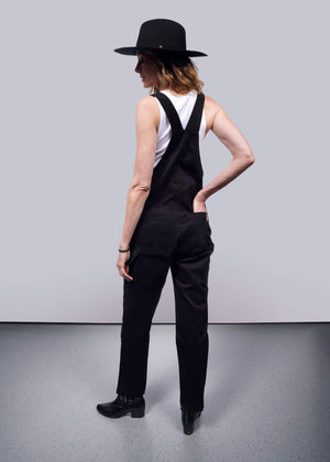 S=Facing away from the camera against a white backdrop, a person sports The Essential Overall in black—a soft stretch cotton design with adjustable straps—paired with a black wide-brimmed hat and white sleeveless top, standing confidently with one hand on their hip.
