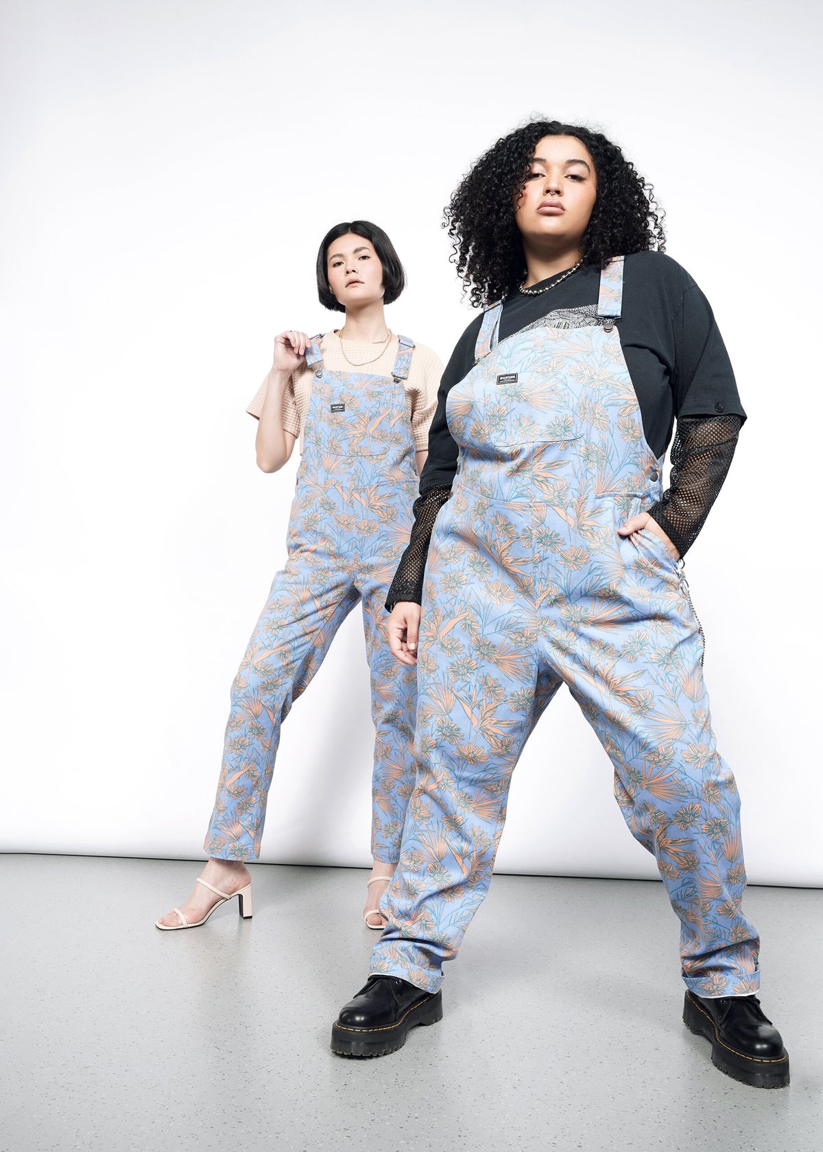 2 Models wearing the Essential Overall in Flower Press Periwinkle. One in size 1X, wearing black mesh long sleeve shirt and black platform shoes. One in size S wearing tan T and tan strappy heeled sandals  