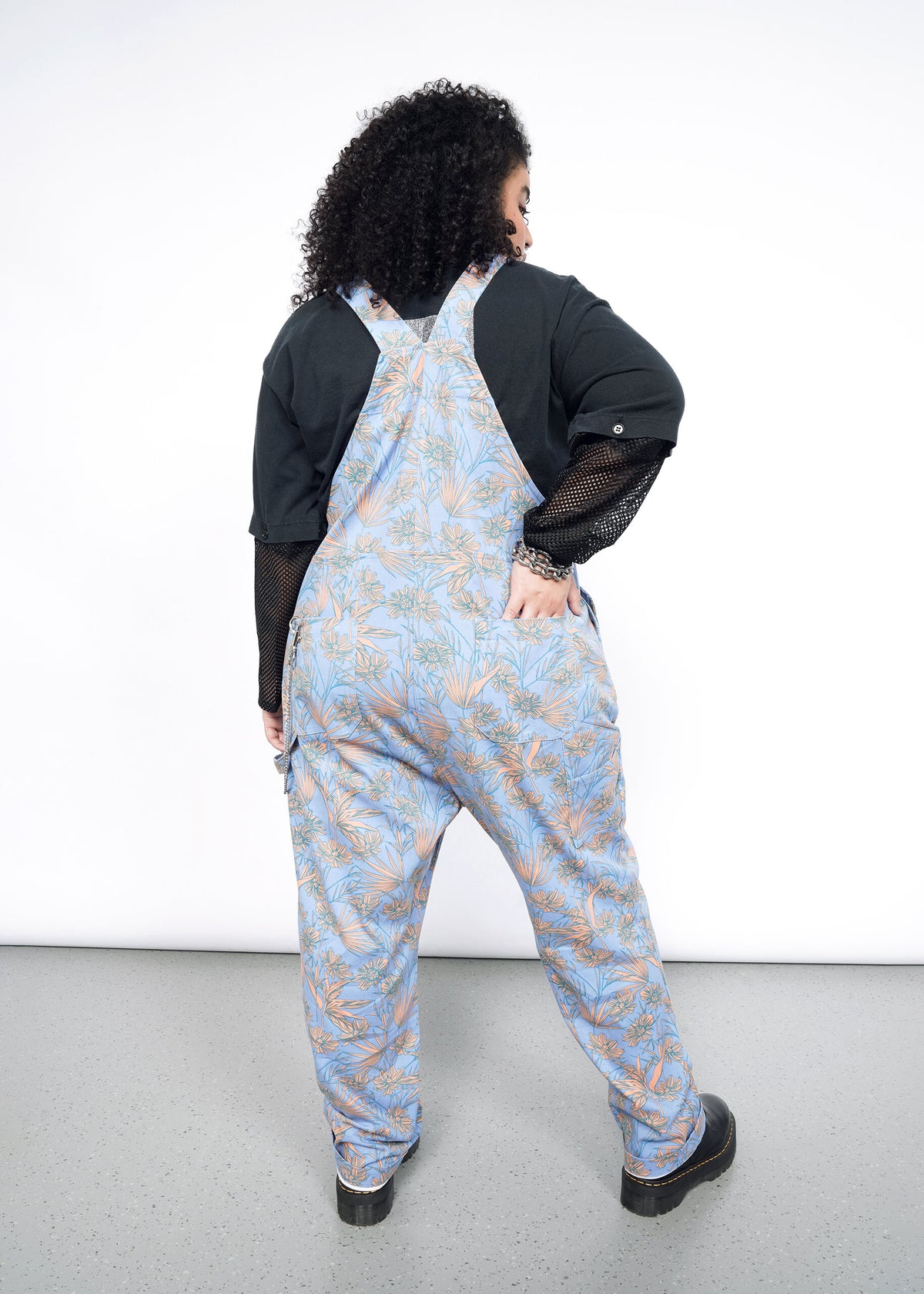 Back image of Model wearing the Essential Overall in Flower Press Periwinkle in size 1X, wearing black mesh long sleeve shirt and black platform shoes.