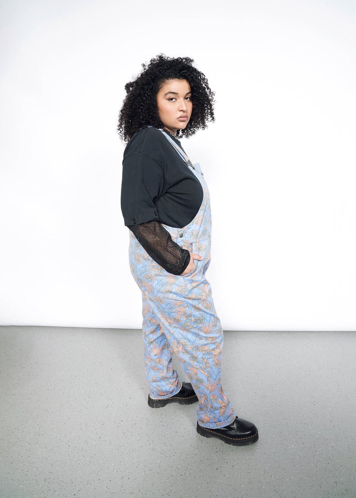 Side image of Model wearing the Essential Overall in Flower Press Periwinkle in size 1X, wearing black mesh long sleeve shirt and black platform shoes.