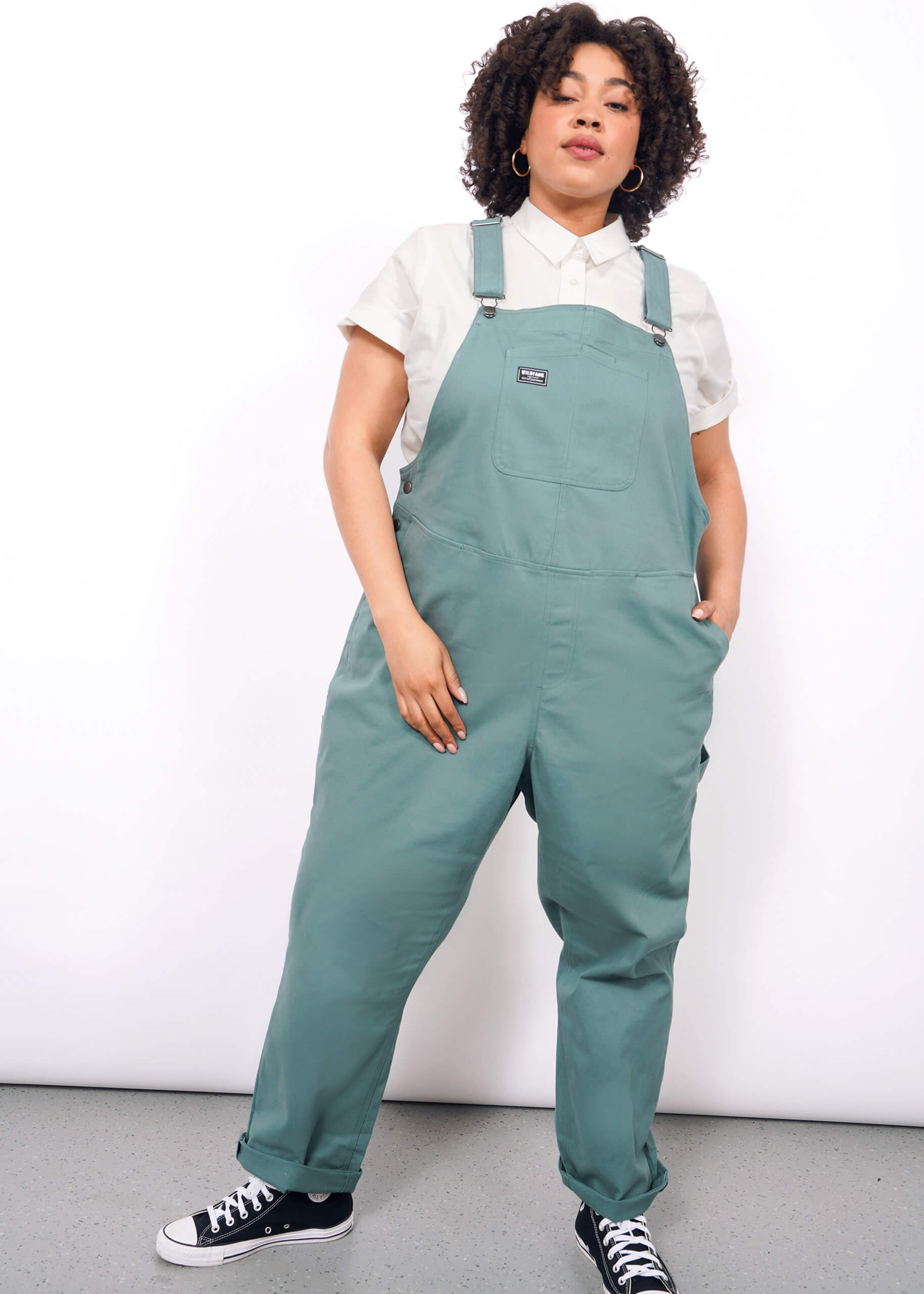 A person with curly hair models The Essential Overall in teal, featuring adjustable straps and deep pockets, styled over a short-sleeved white shirt and black sneakers against a plain white background.