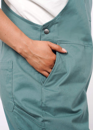 1X=A person wearing The Essential Overall in teal, featuring adjustable straps and a white shirt, has their right hand tucked into the deep front pocket. The image highlights the overalls’ side and the pocket detail.