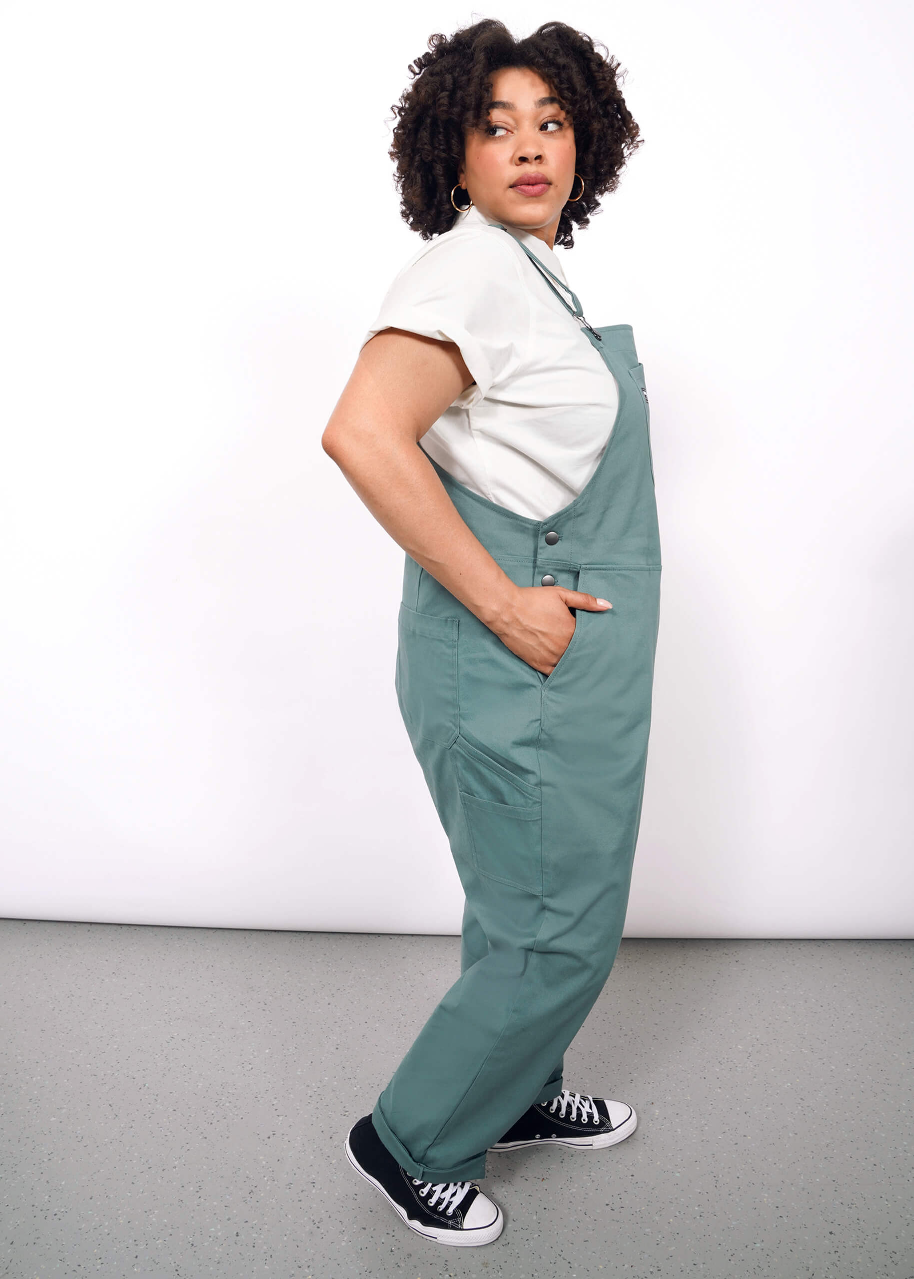 A person with curly hair stands sideways, looking over their shoulder in The Essential Overall—teal, deep-pocketed with adjustable straps—paired with a white t-shirt and black sneakers against a plain white background.