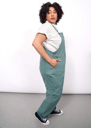1X=A person with curly hair stands sideways, looking over their shoulder in The Essential Overall—teal, deep-pocketed with adjustable straps—paired with a white t-shirt and black sneakers against a plain white background.