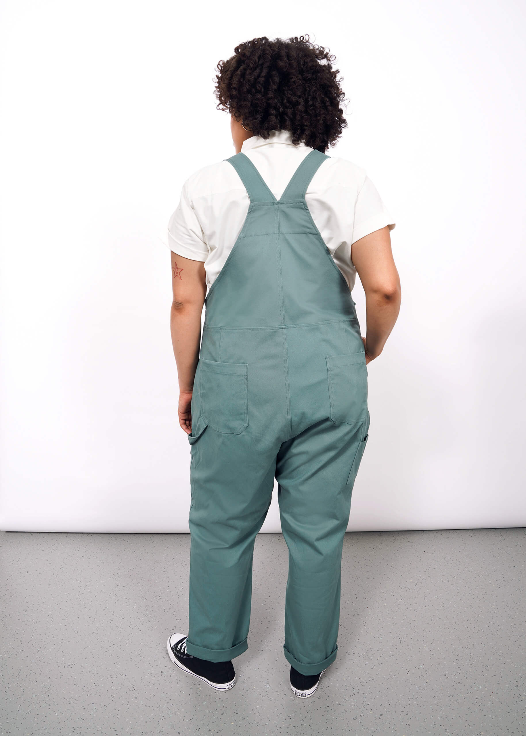 A person with curly hair, wearing The Essential Overall in teal with adjustable straps and a white shirt, stands facing away from the camera against a plain white background.