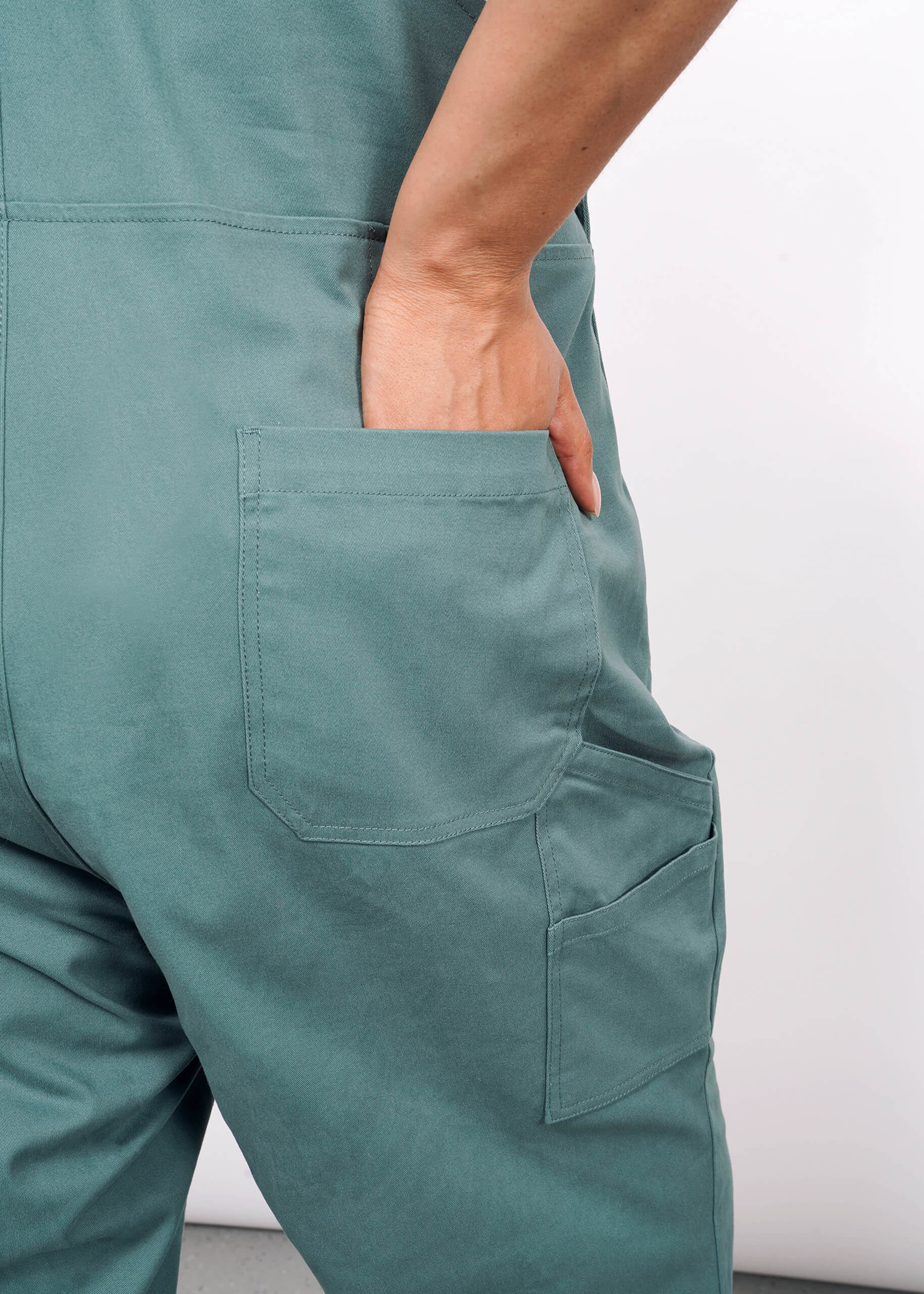 Someone wears The Essential Overall in teal cotton stretch, one hand partly slipped into the deep back pocket, photographed from behind against a plain white background.