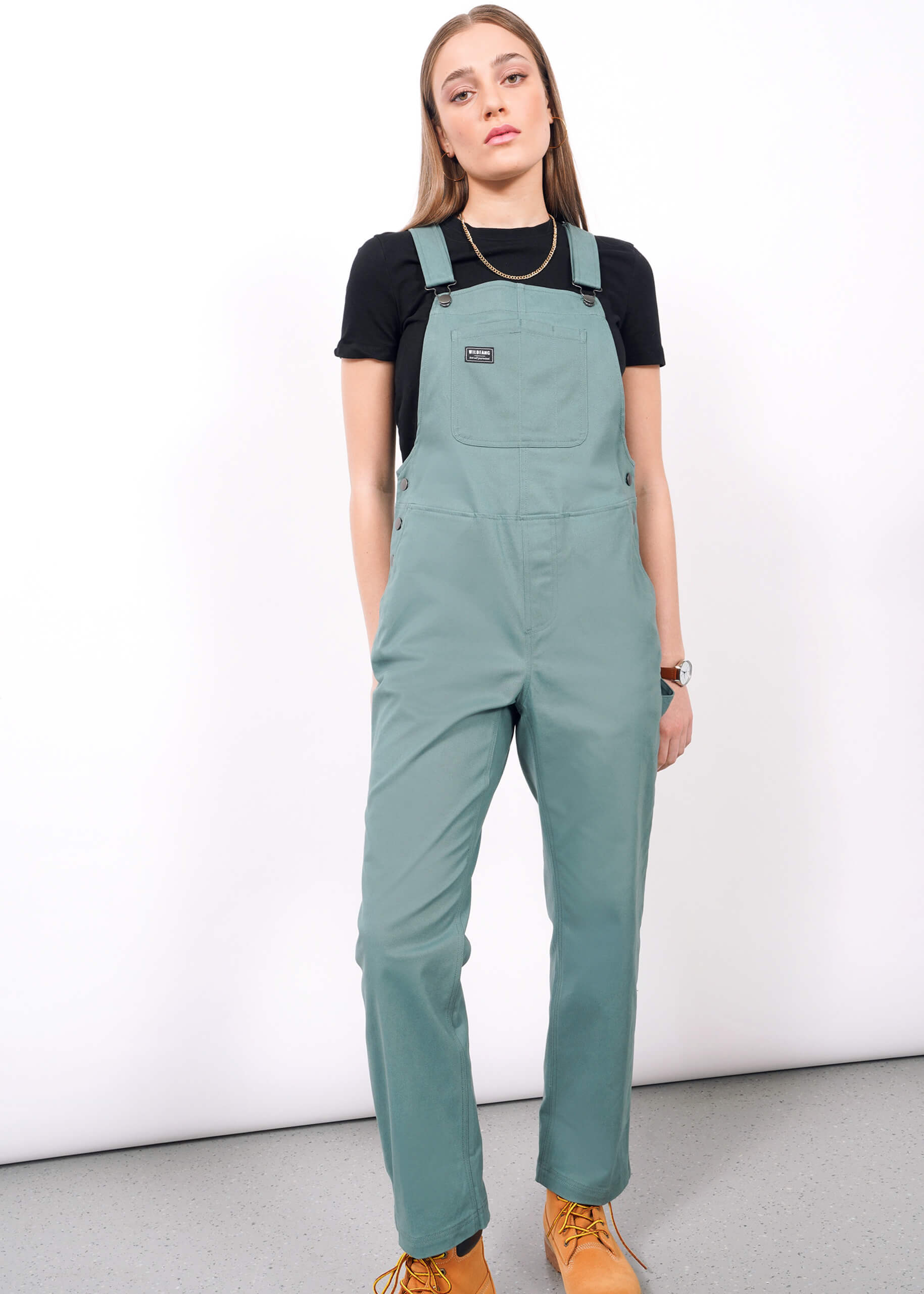 A woman with straight light brown hair and a neutral expression wears The Essential Overall in teal with adjustable straps, paired with a black t-shirt, tan shoes, a necklace, and a wristwatch against a white background.
