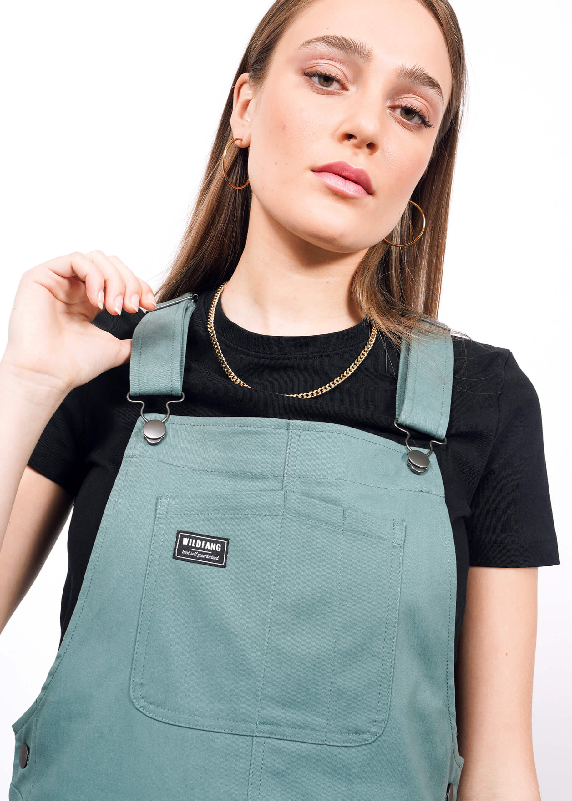 A young woman with straight light brown hair wears gold jewelry, a black t-shirt, and The Essential Overall in green, holding one adjustable strap. She gazes confidently at the camera against a white background.