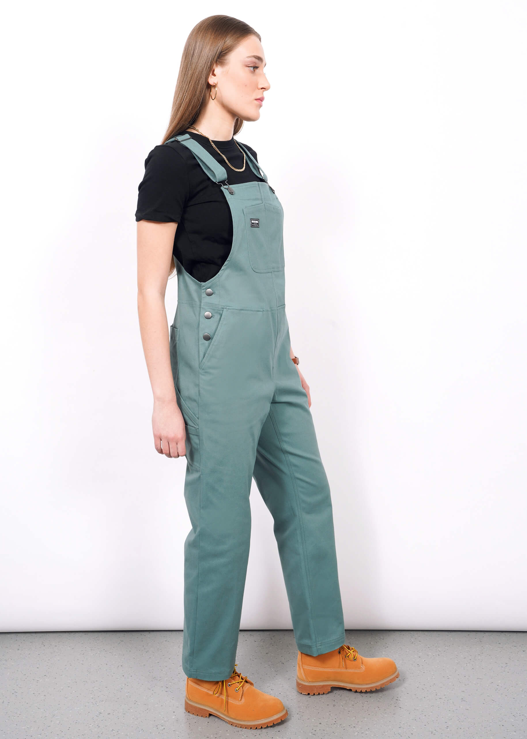 A woman with straight light brown hair and a neutral expression stands sideways against a white background, wearing The Essential Overall in green with adjustable straps and deep pockets, paired with a black t-shirt and tan work boots.