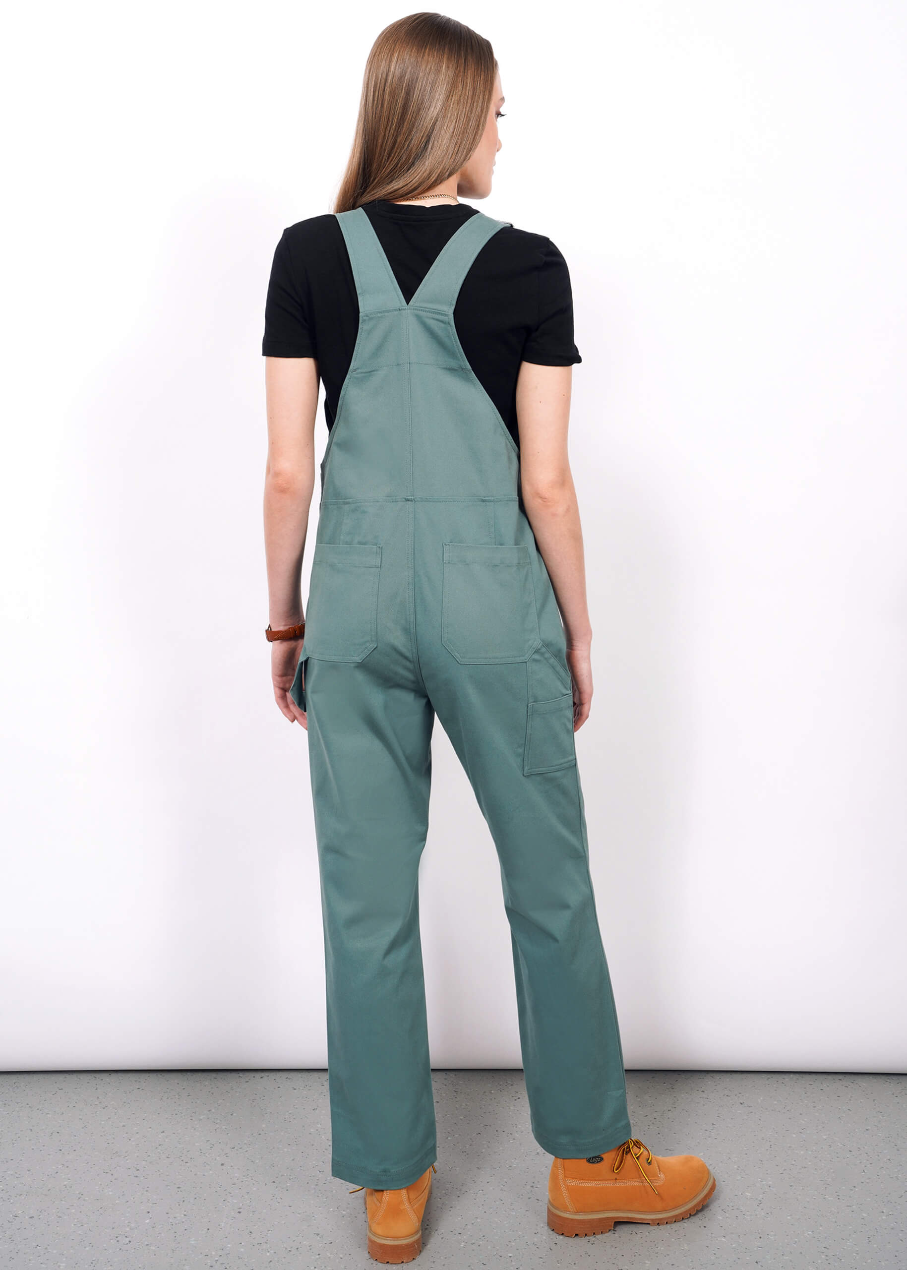 Someone with long hair, wearing The Essential Overall in green with adjustable straps, a black t-shirt, and tan work boots stands facing away from the camera against a plain white wall.