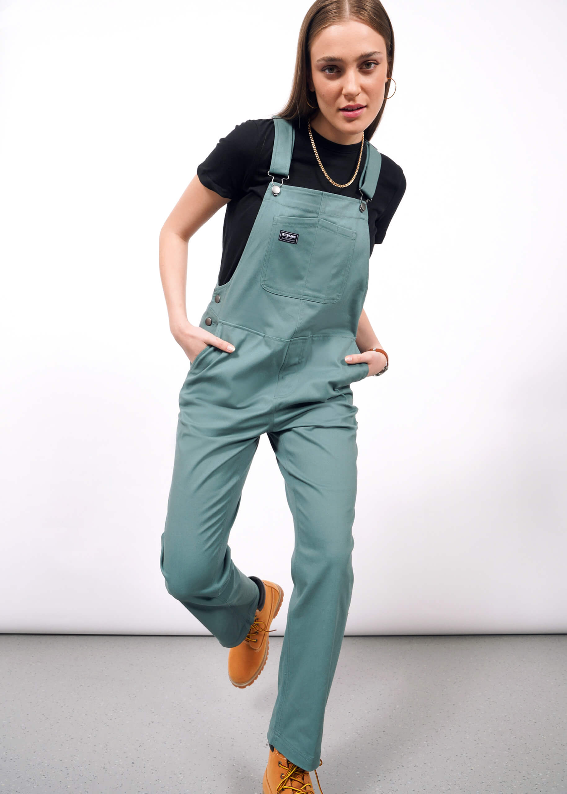 A woman models The Essential Overall in teal—a cotton stretch style with adjustable straps and deep pockets—over a black t-shirt and tan shoes, posing with one leg forward against a plain white background.