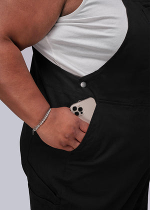 1X=Person wearing black overalls with a white shirt underneath, holding a phone in their pocket.