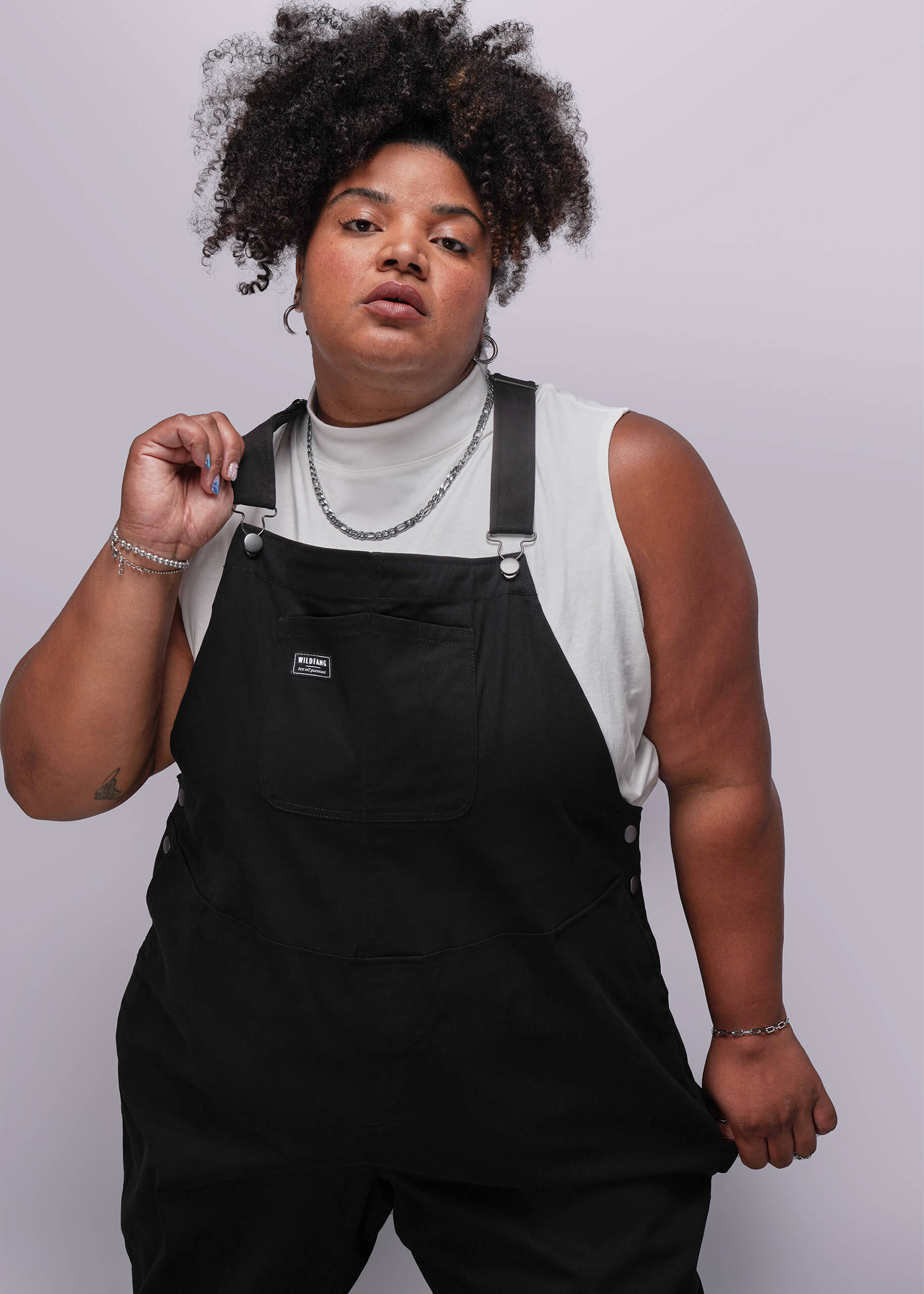 1X=Person wearing black overalls and a light-colored sleeveless top against a plain background