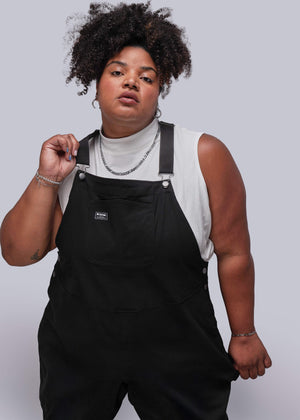 1X=Person wearing black overalls and a light-colored sleeveless top against a plain background