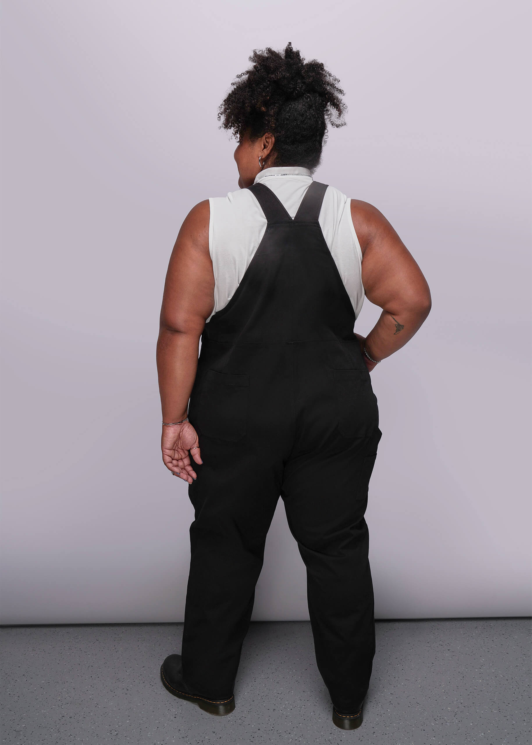1X=Person wearing black overalls with a white shirt underneath against a plain background