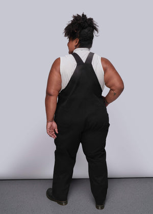 1X=Person wearing black overalls with a white shirt underneath against a plain background