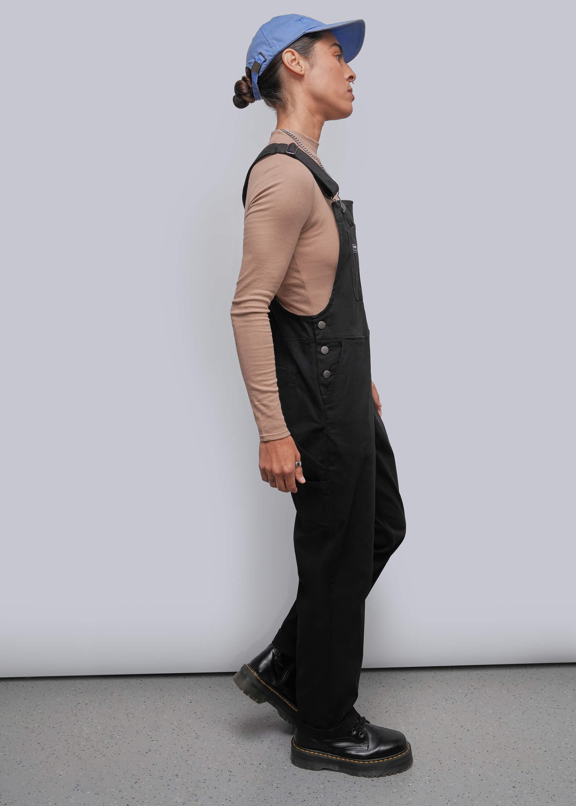 S=A person shown in side profile wearing black overall over a beige long-sleeve top, with a blue baseball cap and black shoes, standing against a plain light gray studio wall and floor.