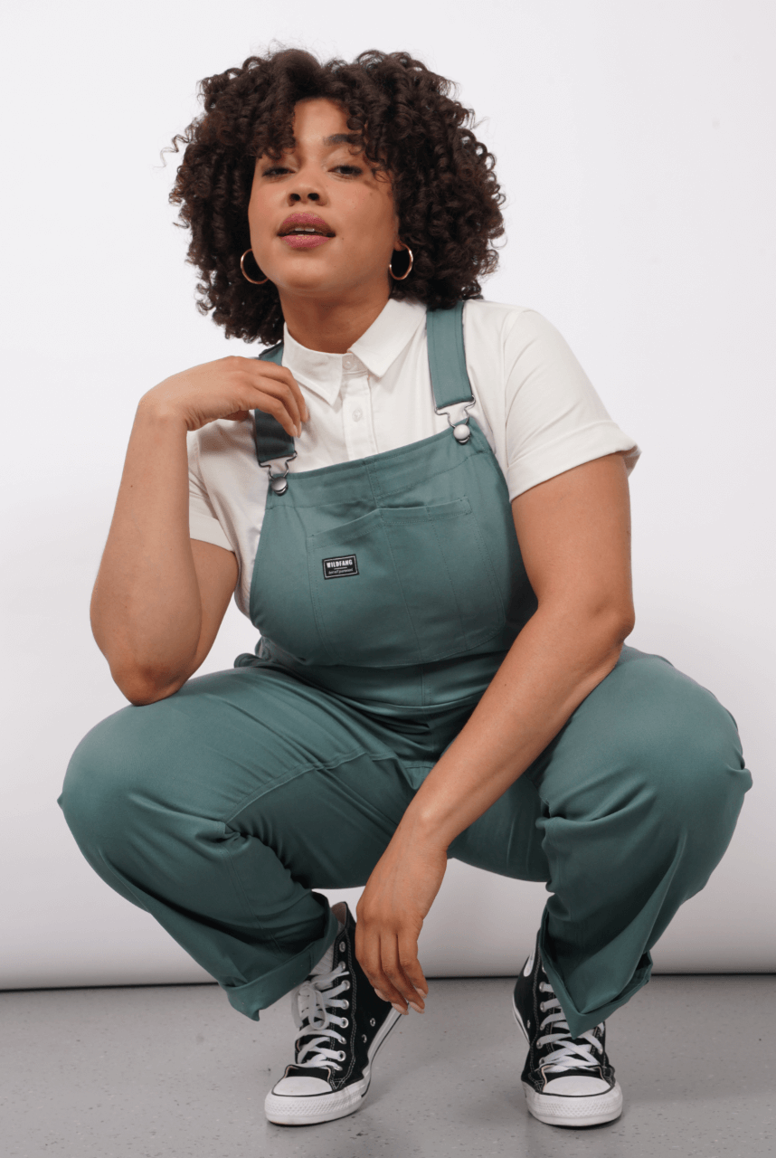 A person with curly hair wearing The Essential Overall—teal, with adjustable straps and deep pockets—over a white button up shirt and paired with black Converse sneakers, squats and looks at camera against a plain white background.