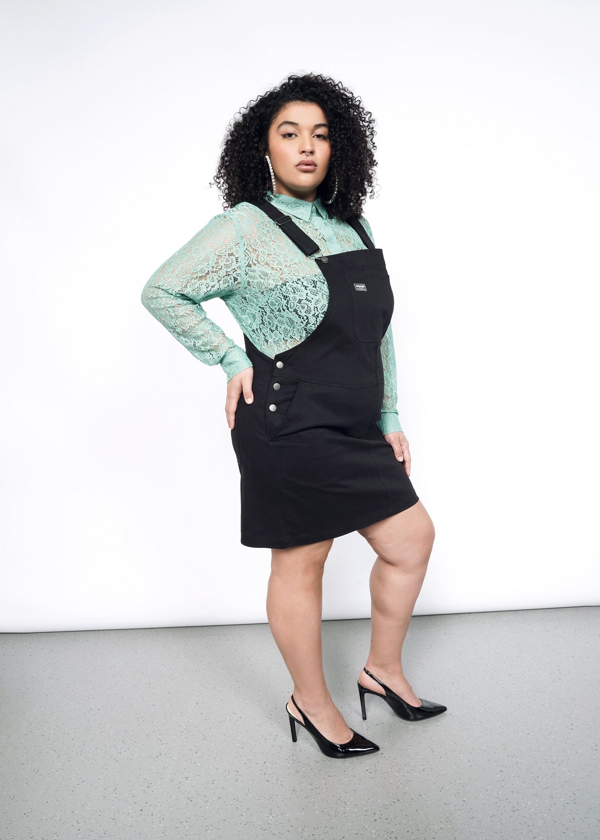 Side image of model wearing the Essential Skirtall in Black size XL, with a Long Sleeve Lace Button Up in Sage and black pumps.