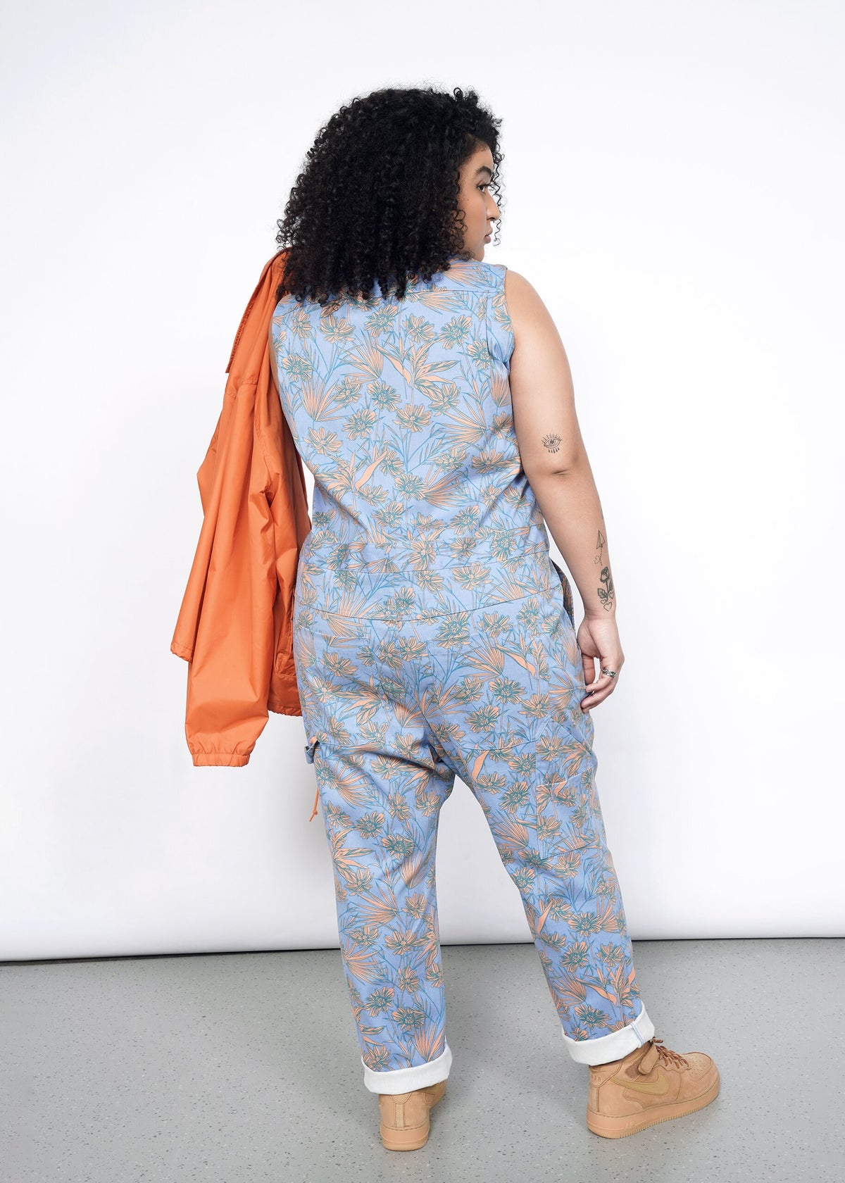 Back image of Model wearing the Essential Sleeveless Coverall in Flower Press Periwinkle in size XL with tan shoes and carrying an orange jacket