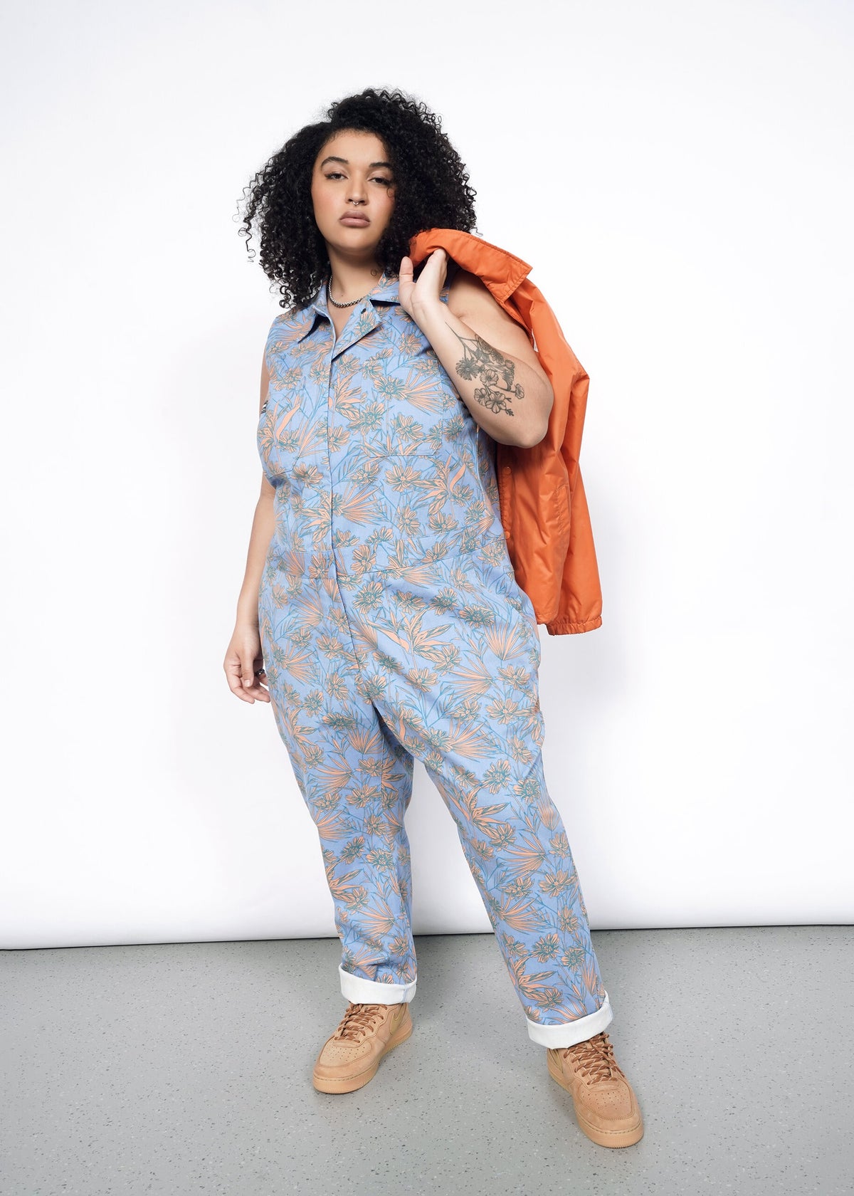 Model wearing the Essential Sleeveless Coverall in Flower Press Periwinkle in size XL with tan shoes and an orange jacket slung over shoulder