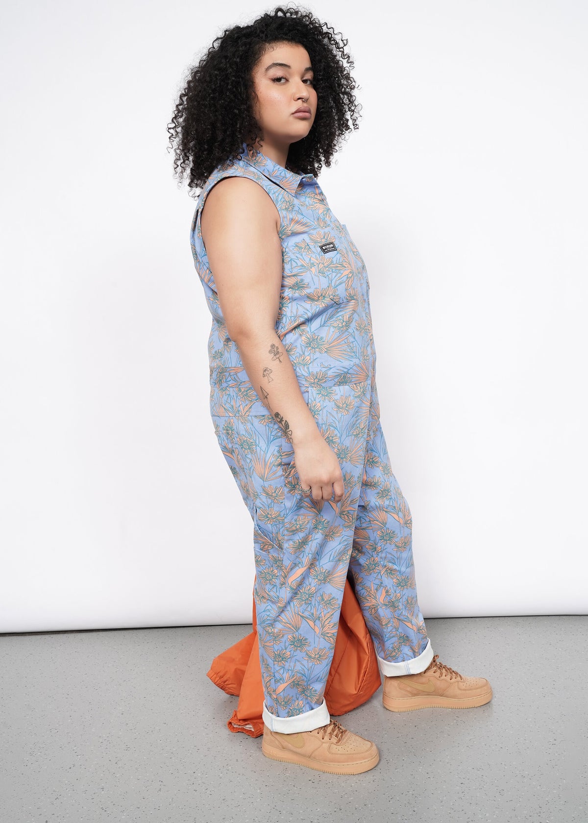 Side image of Model wearing the Essential Sleeveless Coverall in Flower Press Periwinkle in size XL with tan shoes and carrying an orange jacket