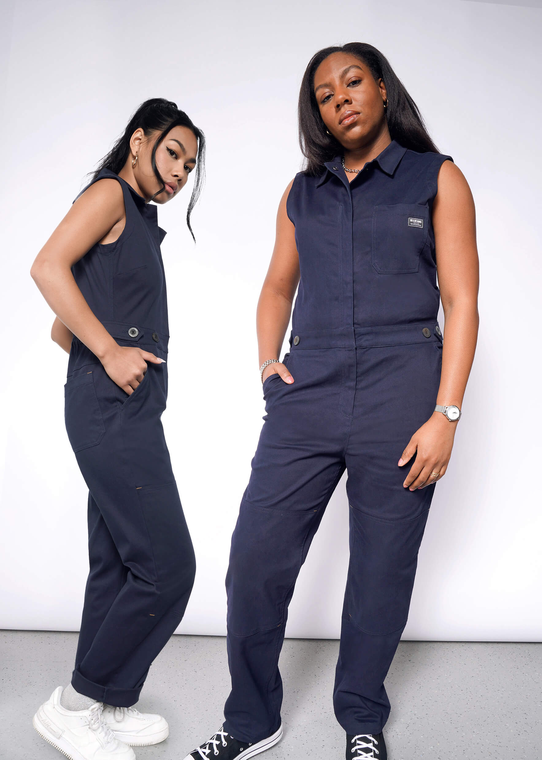 Two women confidently wear The Essential Sleeveless High Waisted Coverall in navy against a white backdrop. The left woman highlights the high-waisted look with one hand on her hip, while the right enjoys its pocketed design and comfortable cotton stretch fabric.