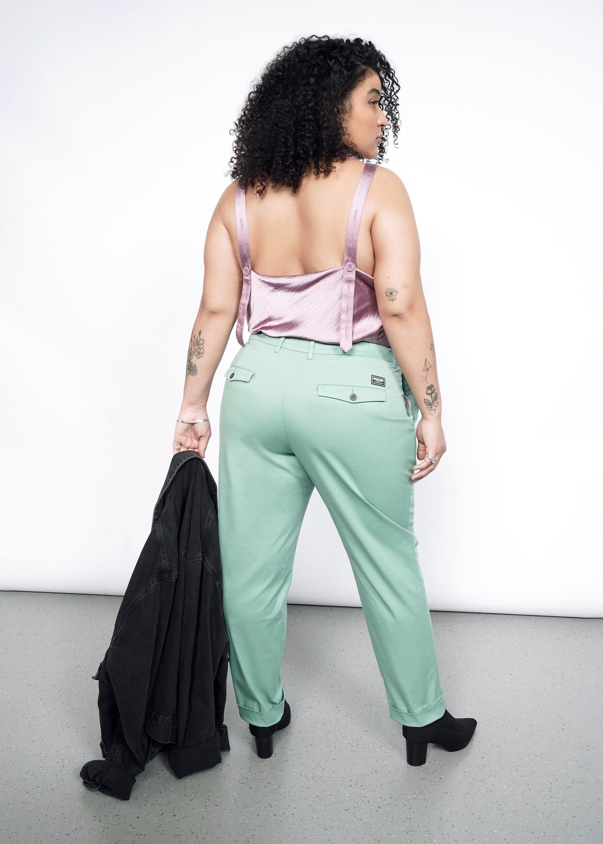 Back image of Model wearing the Essential Trouser in Sage in size 16, with the Empower Cami in Mauve, black heeled boots, and carrying a black jacket.