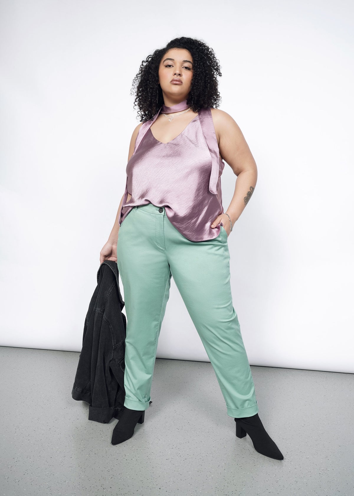 Model wearing the Essential Trouser in Sage in size 16, with the Empower Cami in Mauve, black heeled boots, and carrying a black jacket.