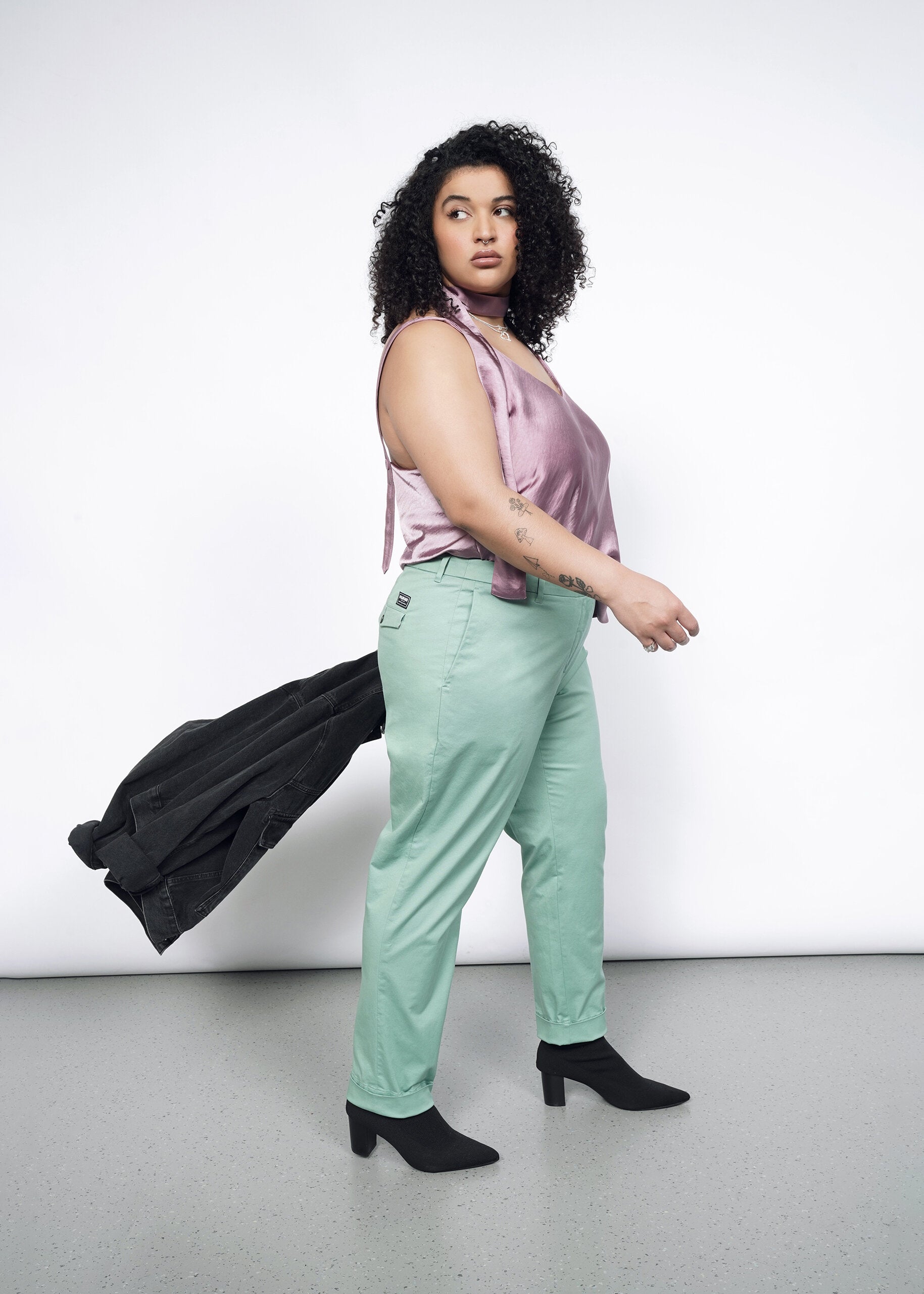 Side image of Model wearing the Essential Trouser in Sage in size 16, with the Empower Cami in Mauve, black heeled boots, and carrying a black jacket.