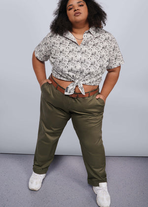 1X=A curly-haired person confidently stands with hands in pockets, wearing a patterned short-sleeve shirt tied at the waist. They pair The Essential Trouser in olive green, a wardrobe staple, with a brown belt and white sneakers against a gray and white gradient background.