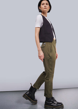 S= Against a plain backdrop, a person sports The Essential Trouser in olive green, paired with a white t-shirt, black vest, and black boots. They have short dark hair and glance sideways.