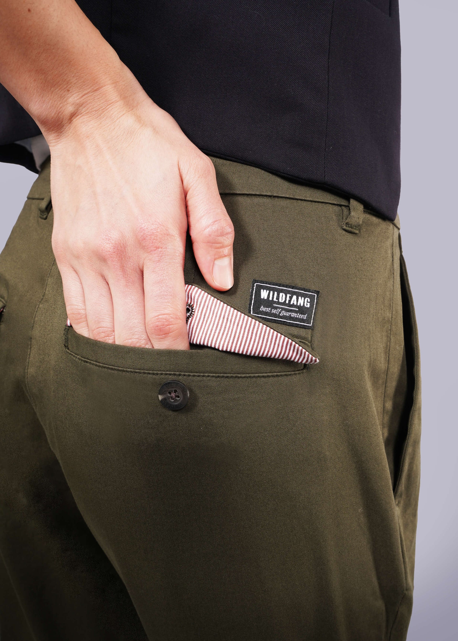 S=Someone wearing The Essential Trouser in olive-green—a wardrobe staple with a WILDFANG label—tucks a striped handkerchief into the back pocket. Their hand, part of the pocket, and a black shirt are also visible.