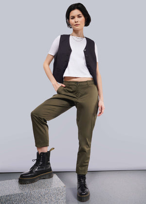 S=A person with short dark hair poses confidently in a white t-shirt, a black vest, and The Essential Trouser in olive green. These wardrobe staples pair seamlessly with their black boots as they stand with one hand in their pocket against a neutral background.