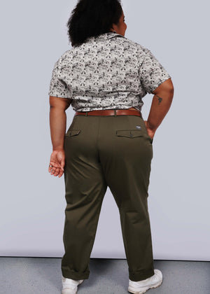 1X= Against a white backdrop, a person faces away, wearing a patterned shirt with convertible cuffs and The Essential Trouser in olive green, belt-cinched as a wardrobe classic. Their feet are adorned with white sneakers, and their right forearm features an intriguing tattoo.