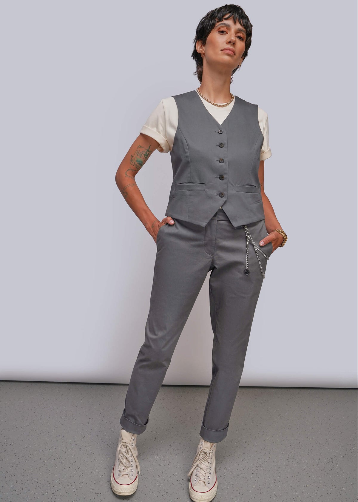 S=A model wearing a gray vest and matching gray trousers over a white t-shirt, styled with white sneakers, standing against a light gray studio background.
