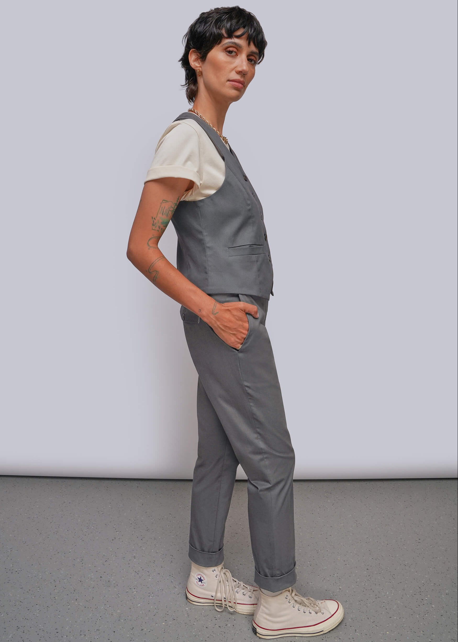 A model wearing a gray vest over a cream t-shirt, gray pants, and off-white high-top sneakers, standing against a light gray studio background.