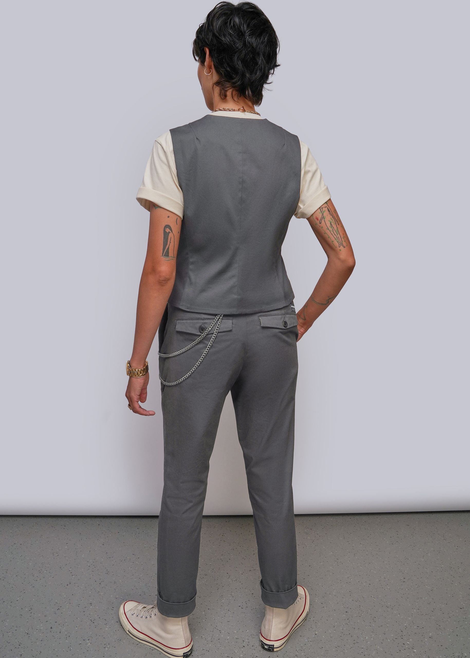 S=A model shown from the back wearing a dark gray sleeveless vest over a cream t-shirt, dark gray trousers with a chain detail at the back pocket, and beige sneakers, set against a plain light gray studio background.
