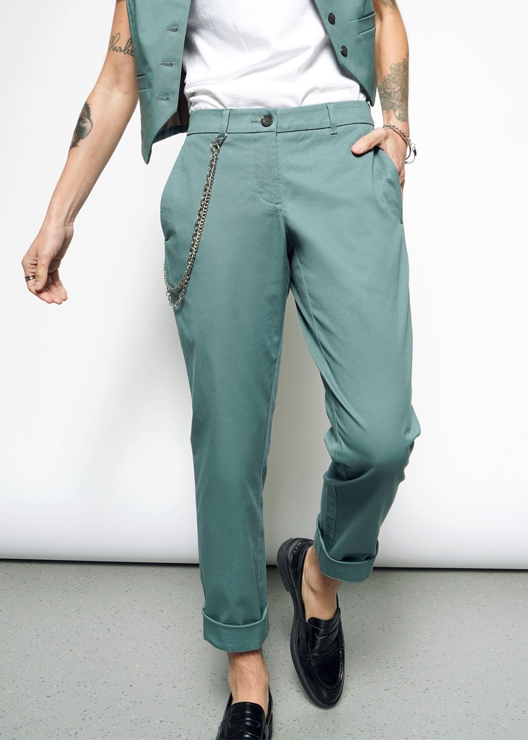 A person stands casually with a tattooed arm, hand in their pocket, wearing The Essential Trouser in classic teal with a chain accessory, paired with a white shirt and cropped teal vest. They wear black loafers against a plain white background.