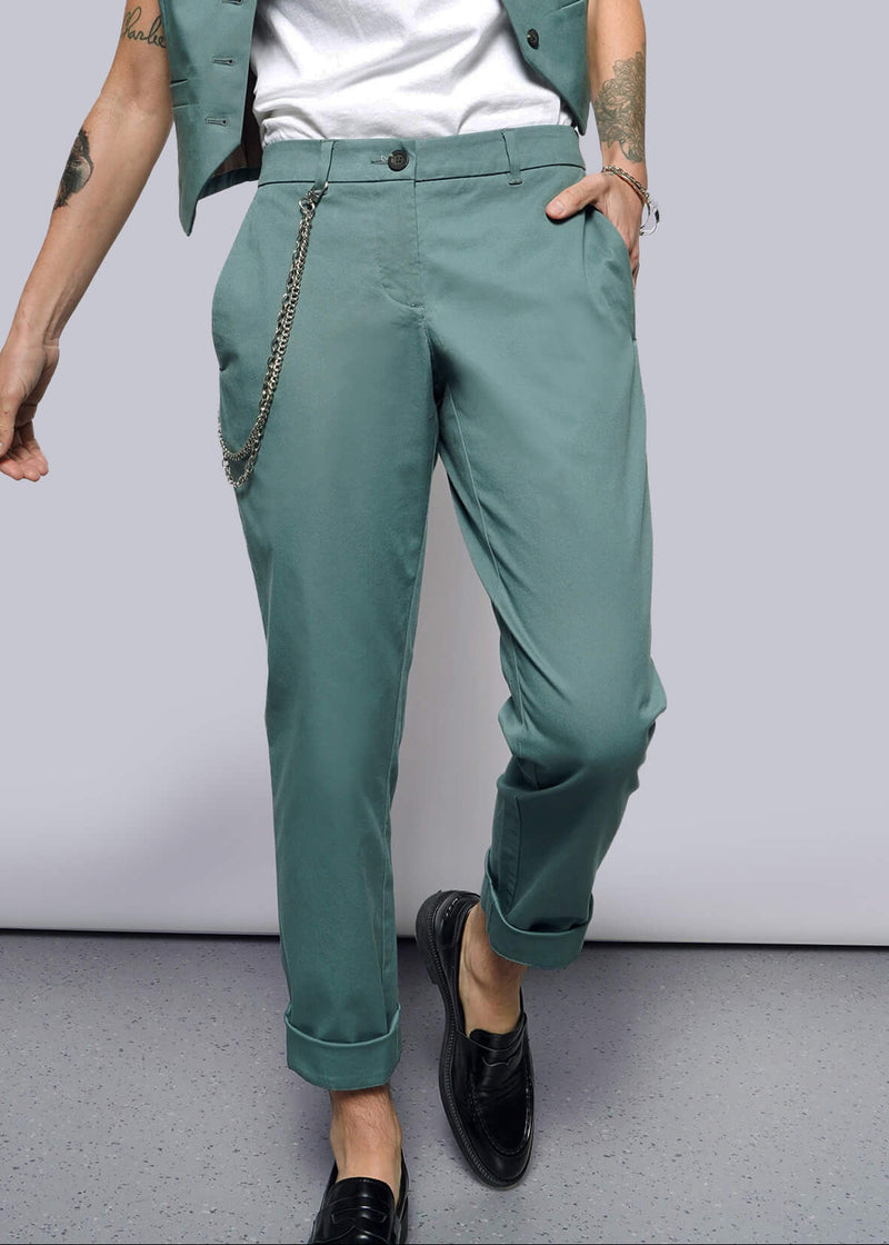 S= A person stands casually with a tattooed arm, hand in their pocket, wearing The Essential Trouser in classic teal with a chain accessory, paired with a white shirt and cropped teal vest. They wear black loafers against a plain white background.