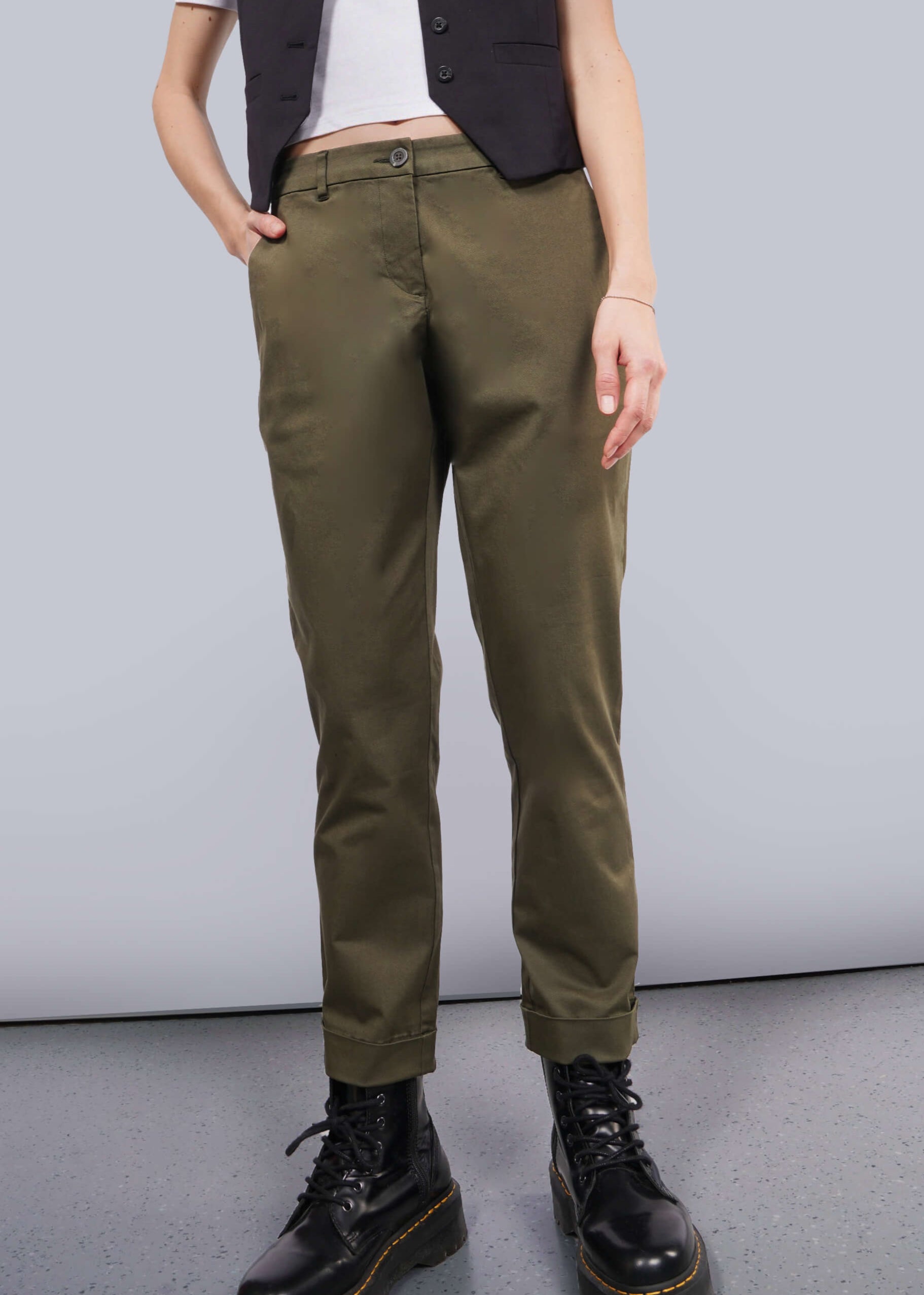 Someone wearing The Essential Trouser in olive green, perfectly paired with a black sleeveless vest and sleek black boots. Their hands rest casually in the pockets against a plain white background that enhances the minimalist aesthetic.